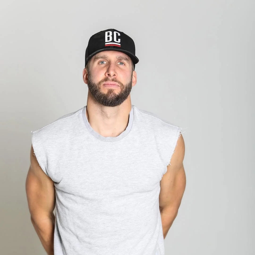 A man with a beard and blue eyes wearing a black cap with 'BC' on it and a sleeveless gray shirt, standing against a plain light gray background.