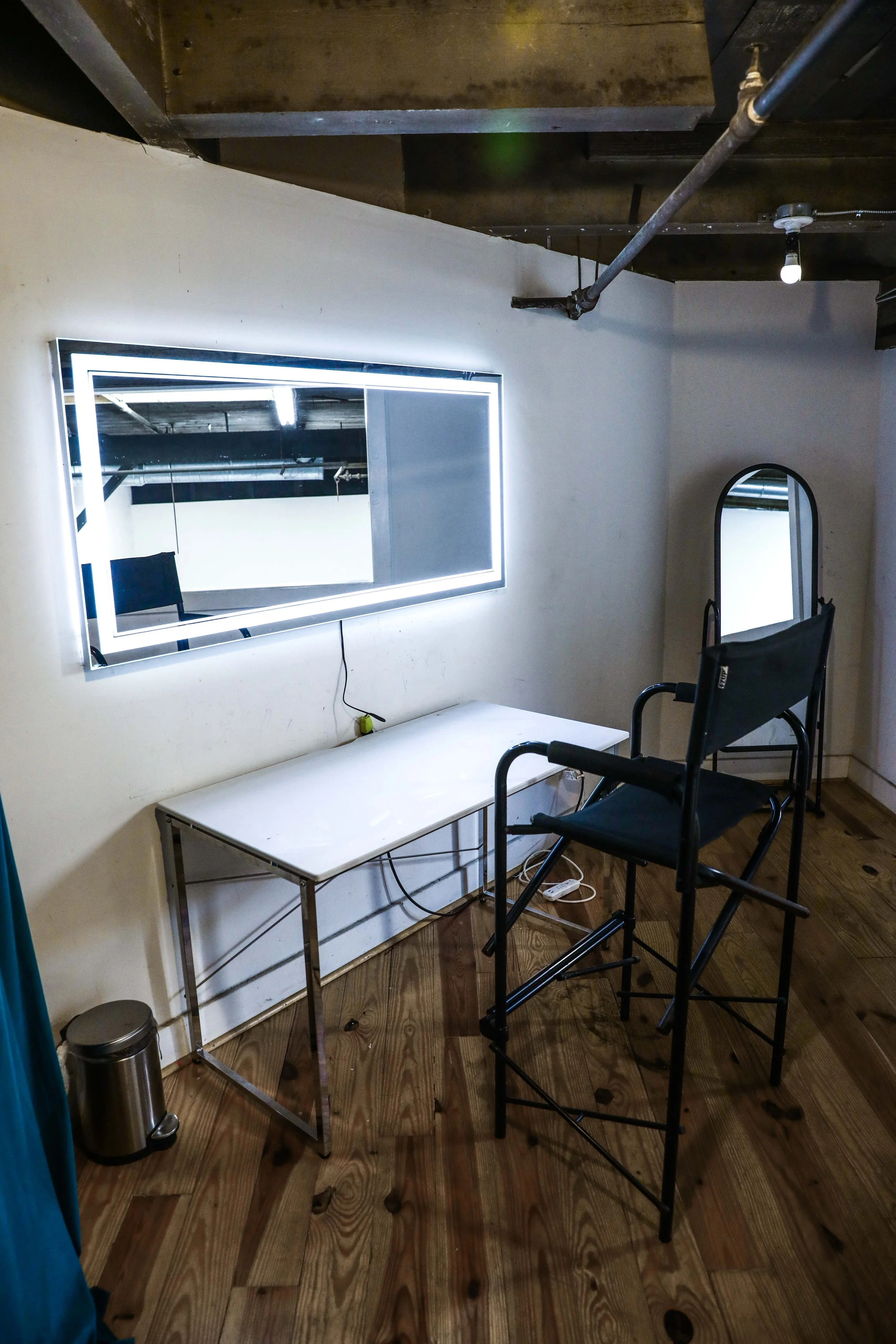 A room with a white wall, wooden floor, and exposed ceiling. There is a illuminated rectangular mirror mounted on the wall, a white table with a metal frame underneath, a black chair with a high backrest, a tall mirror with a black frame, and a small