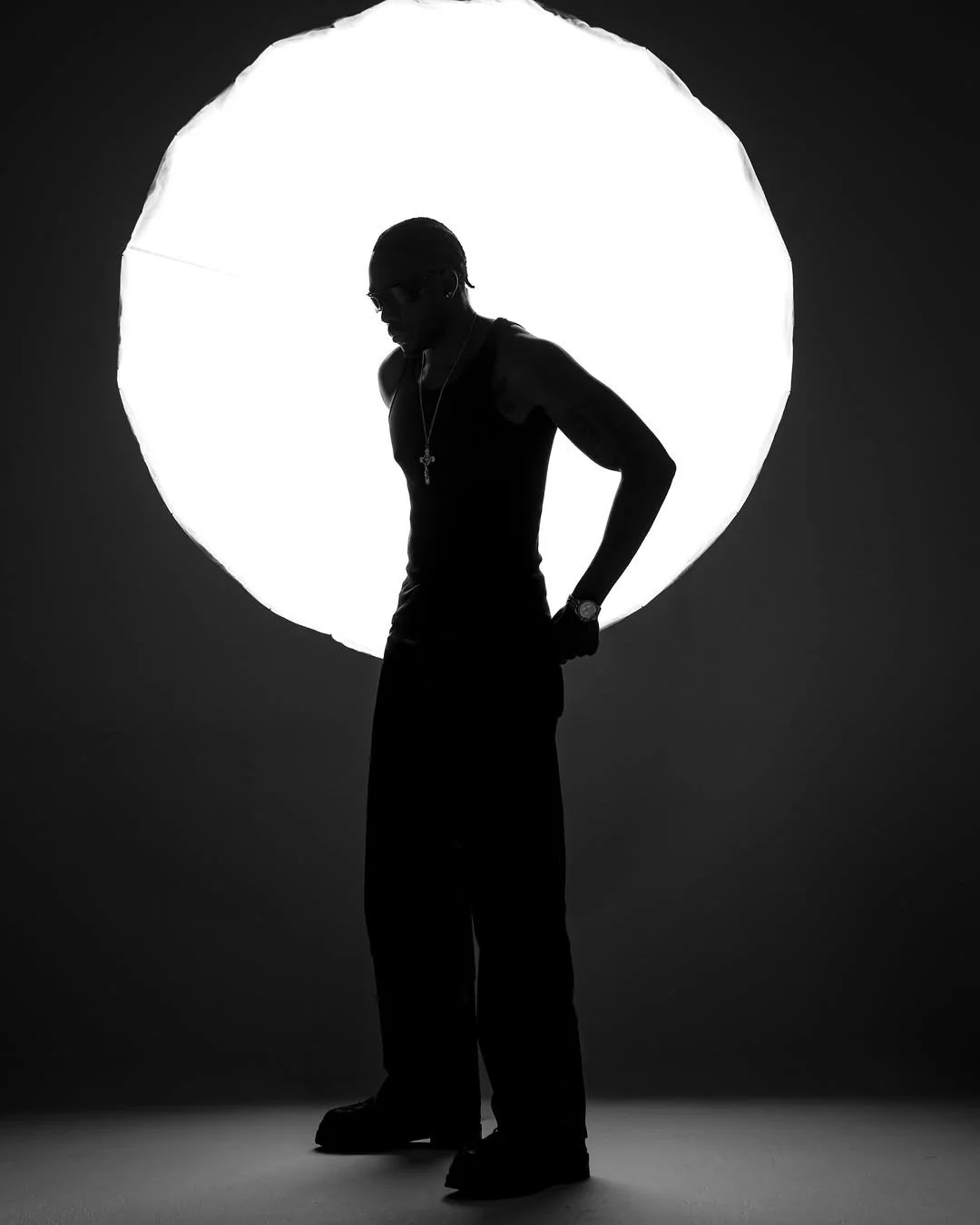 Silhouette of a man wearing a cap, sunglasses, a watch, a necklace with a cross, and earrings, standing in front of a large bright circular light source.