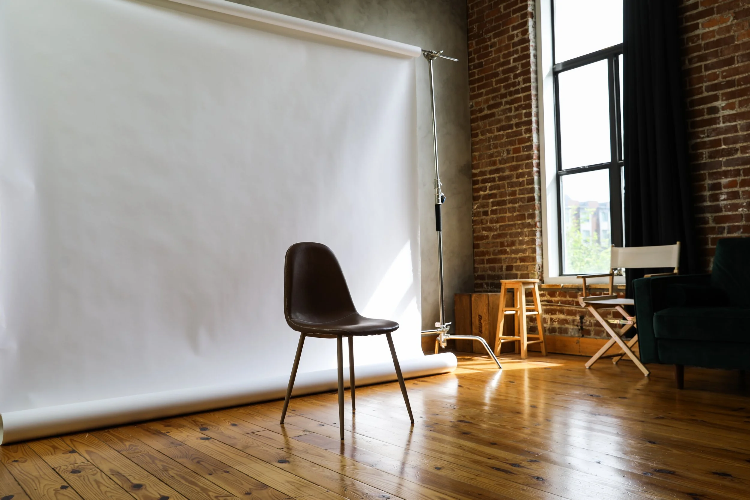 An indoor photography studio with a white backdrop, a black chair, a wooden stool, a director's chair, and a sofa by a large window with brick walls and wooden floors.
