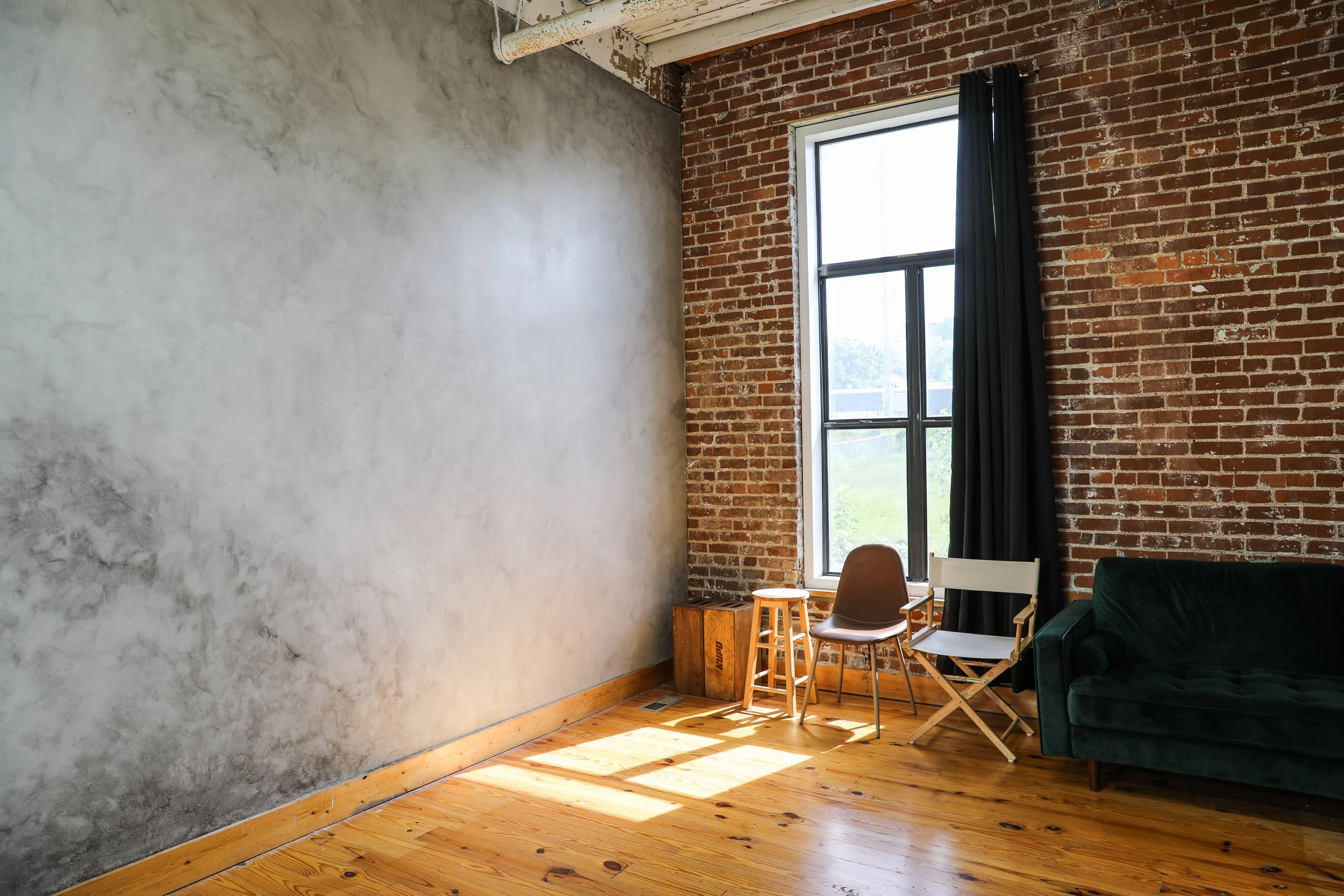 Interior of an industrial-style room with a brick wall, large window with black curtain, hardwood floor, and a green velvet couch, along with chairs, a wooden stool, and a small wooden box.