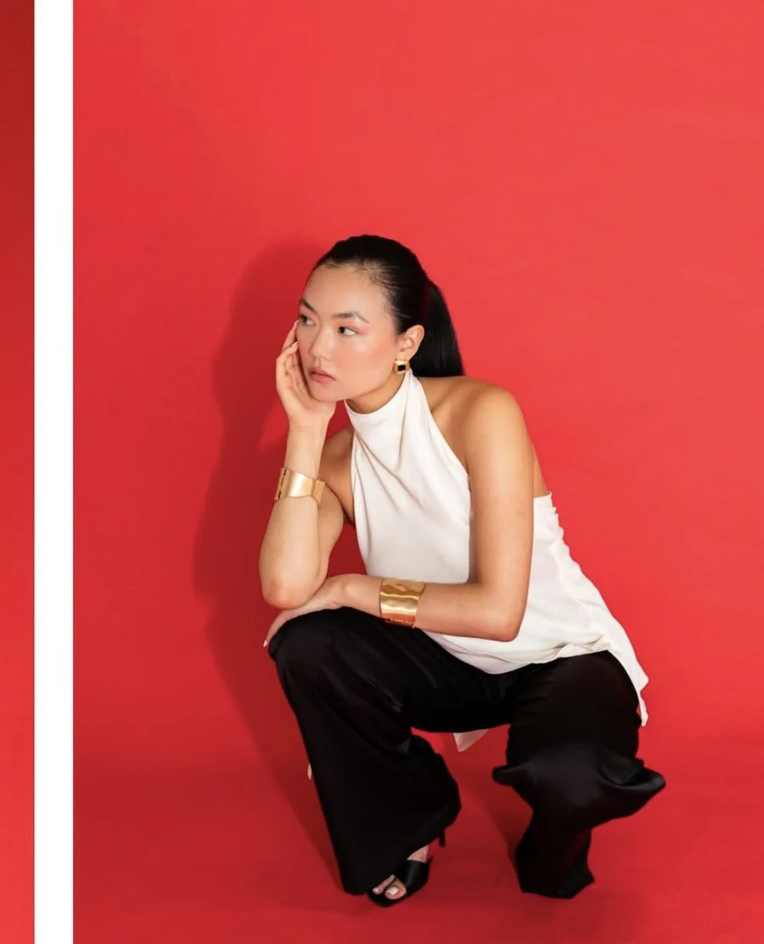 A woman with black hair dressed in a white sleeveless top and black pants, posing against a red background, wearing gold jewelry.