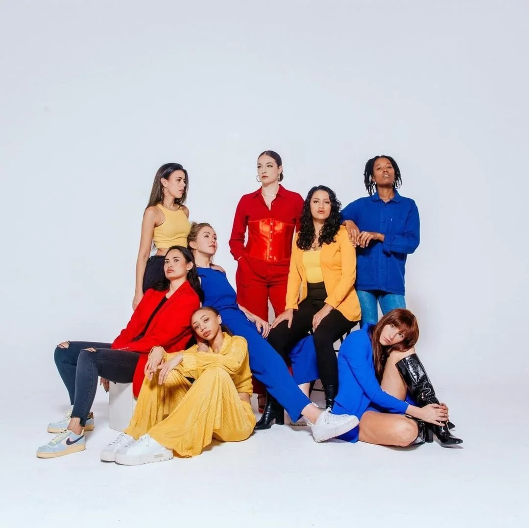 Group of nine diverse women posing together against a plain white background, wearing colorful outfits including red, blue, yellow, and black.