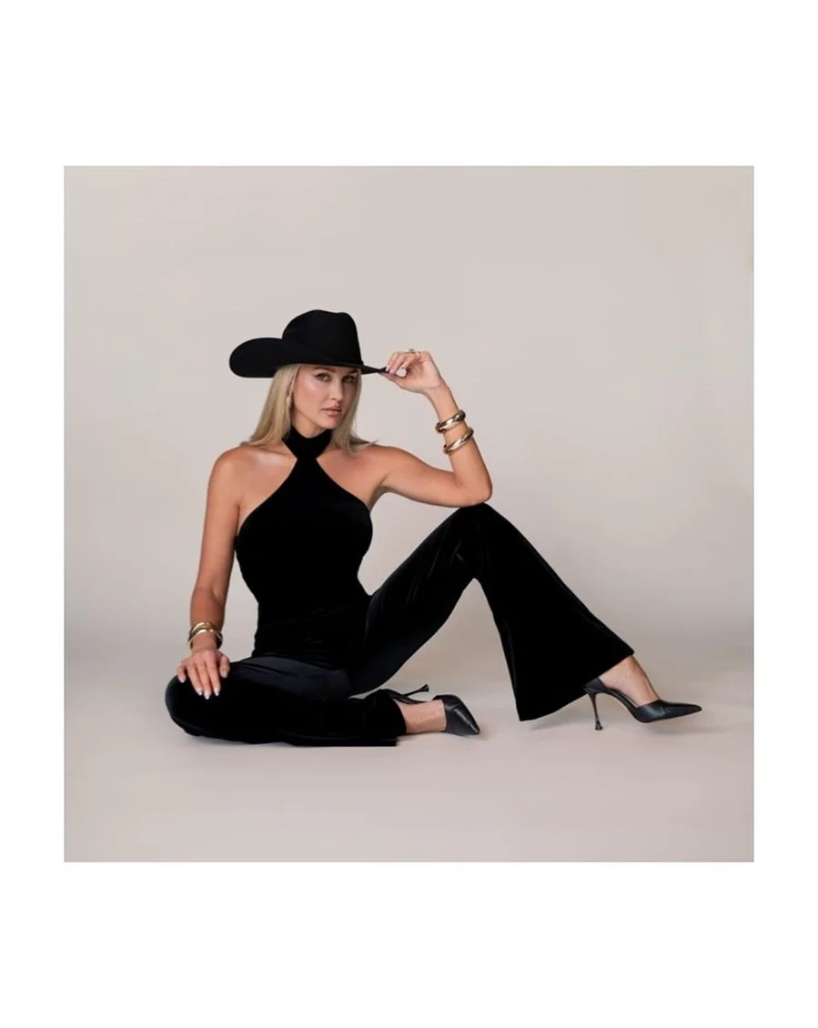 A woman wearing a black wide-brimmed hat, black halter top, black velvet pants, and black high heels, sitting on the floor against a plain neutral background. She has blonde hair, jewelry including bracelets, and is holding the brim of her hat with o