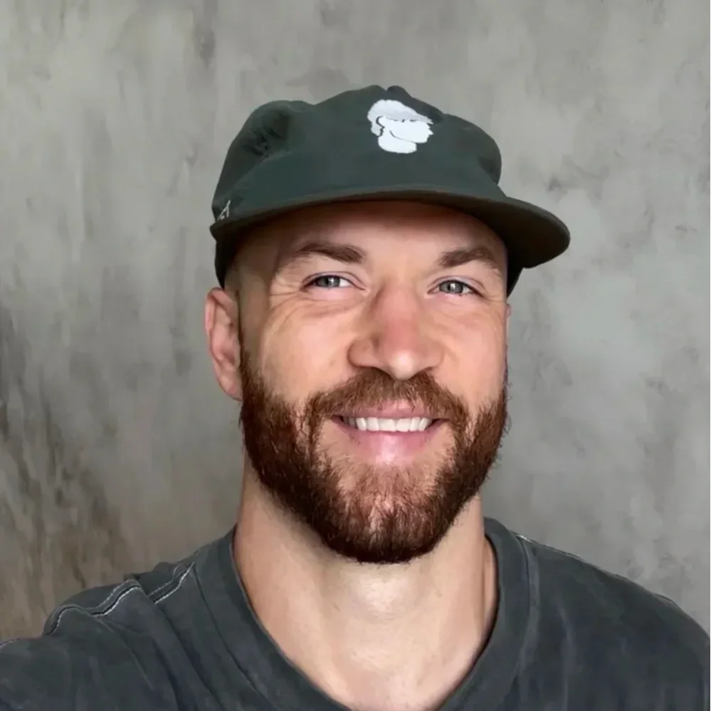 Close-up of a smiling man with a beard wearing a black cap with a white logo and a dark gray shirt, standing in front of a gray concrete wall.