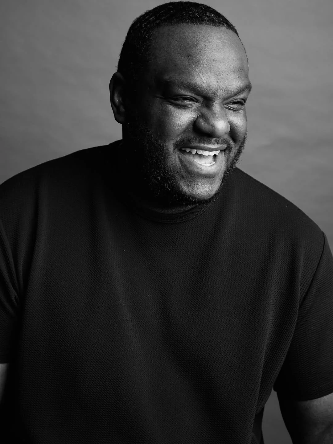 A black and white portrait of a man smiling, wearing a dark t-shirt.