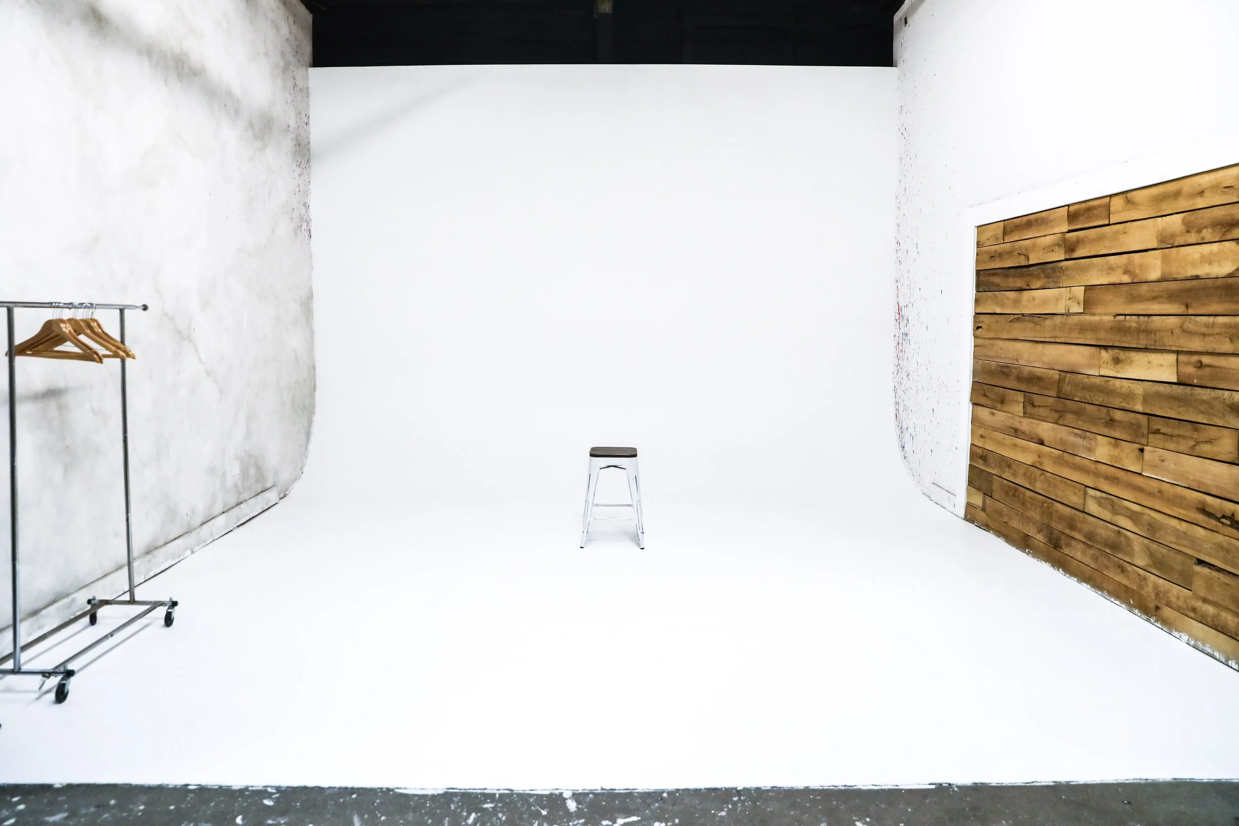 A minimalist photography or video studio with a white arched backdrop, a stool in the center, a black clothing rack with a couple of wooden hangers on the left, a textured white wall on the left, and a wooden-paneled wall on the right.