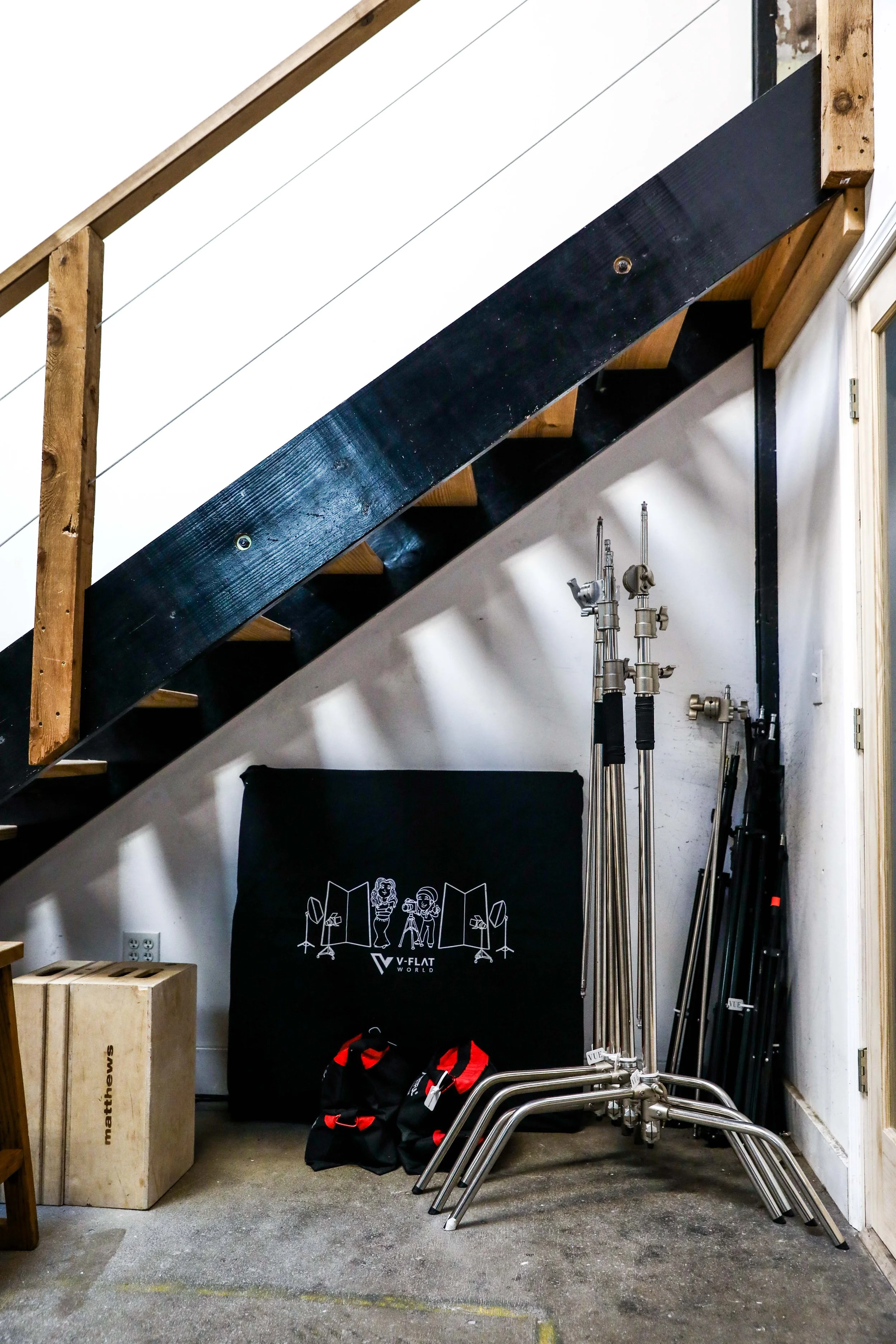 Photograph of a storage area under a staircase with photography equipment, including lighting stands, bags, a black backdrop with a logo, and a wooden box labeled 'mathews'.