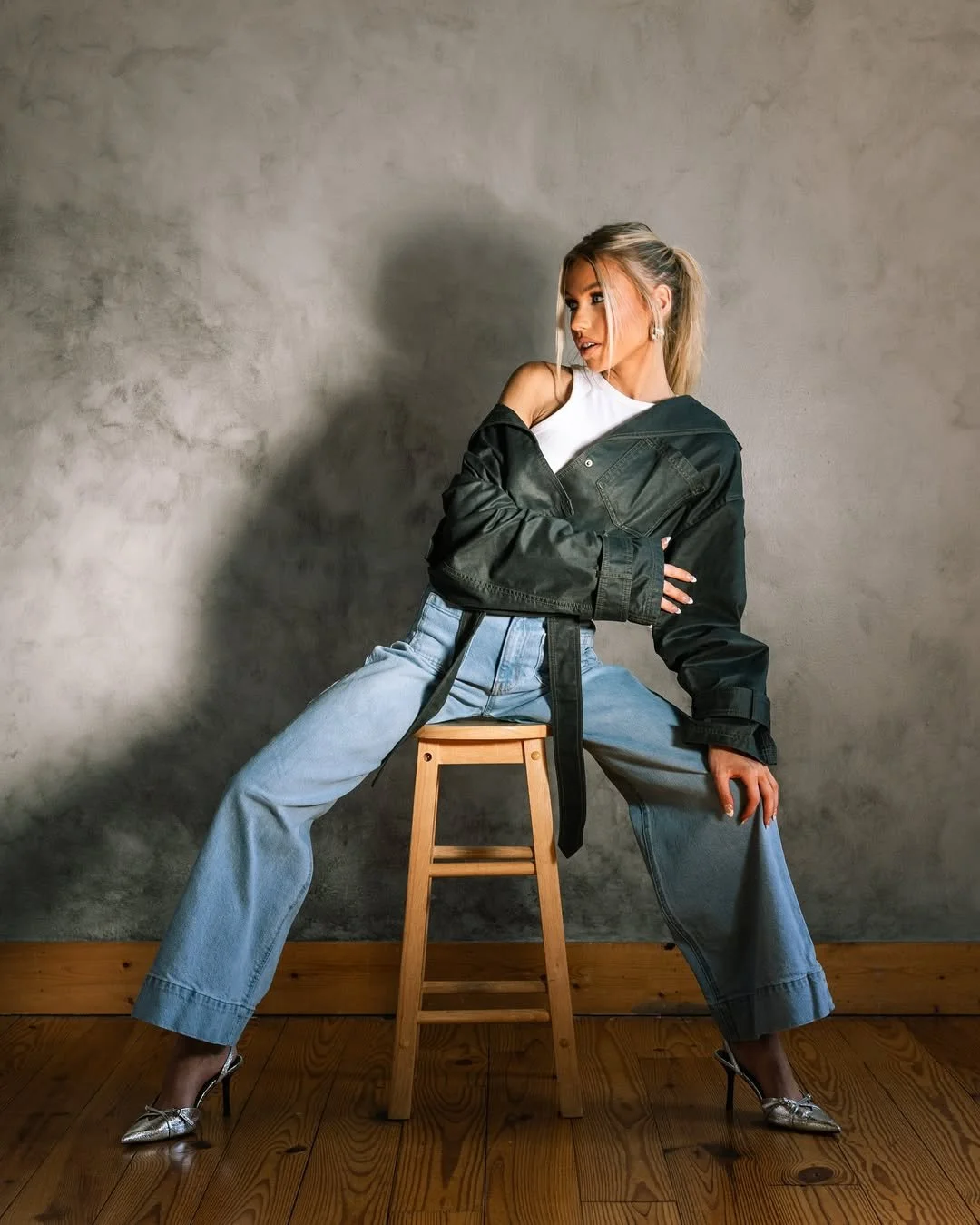 A woman wearing jeans, silver heels, a white top, and a black jacket draped over her shoulders, sitting on a wooden stool against a textured gray wall, striking a pose with her legs wide apart and looking to her right.