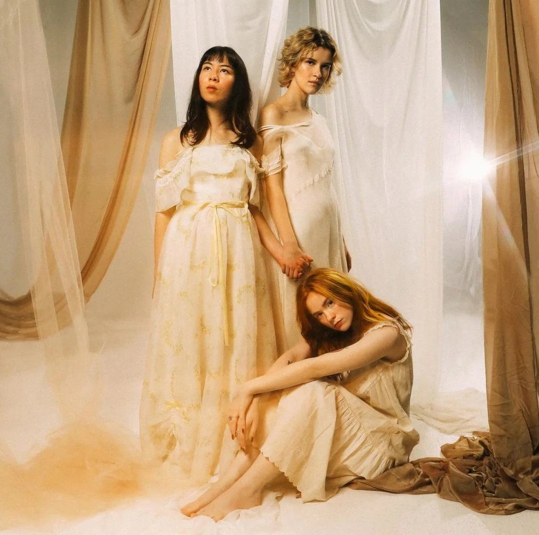 Three women in cream-colored dresses in a softly lit room with sheer curtains. One woman is sitting on the floor, two are standing and holding hands.