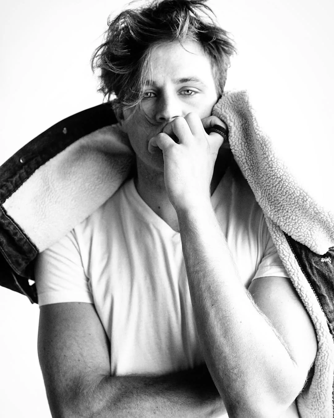 Black and white photo of a young person with tousled hair, wearing a white t-shirt, holding a fleece-lined jacket over their shoulders, one hand near their face, looking directly at the camera.