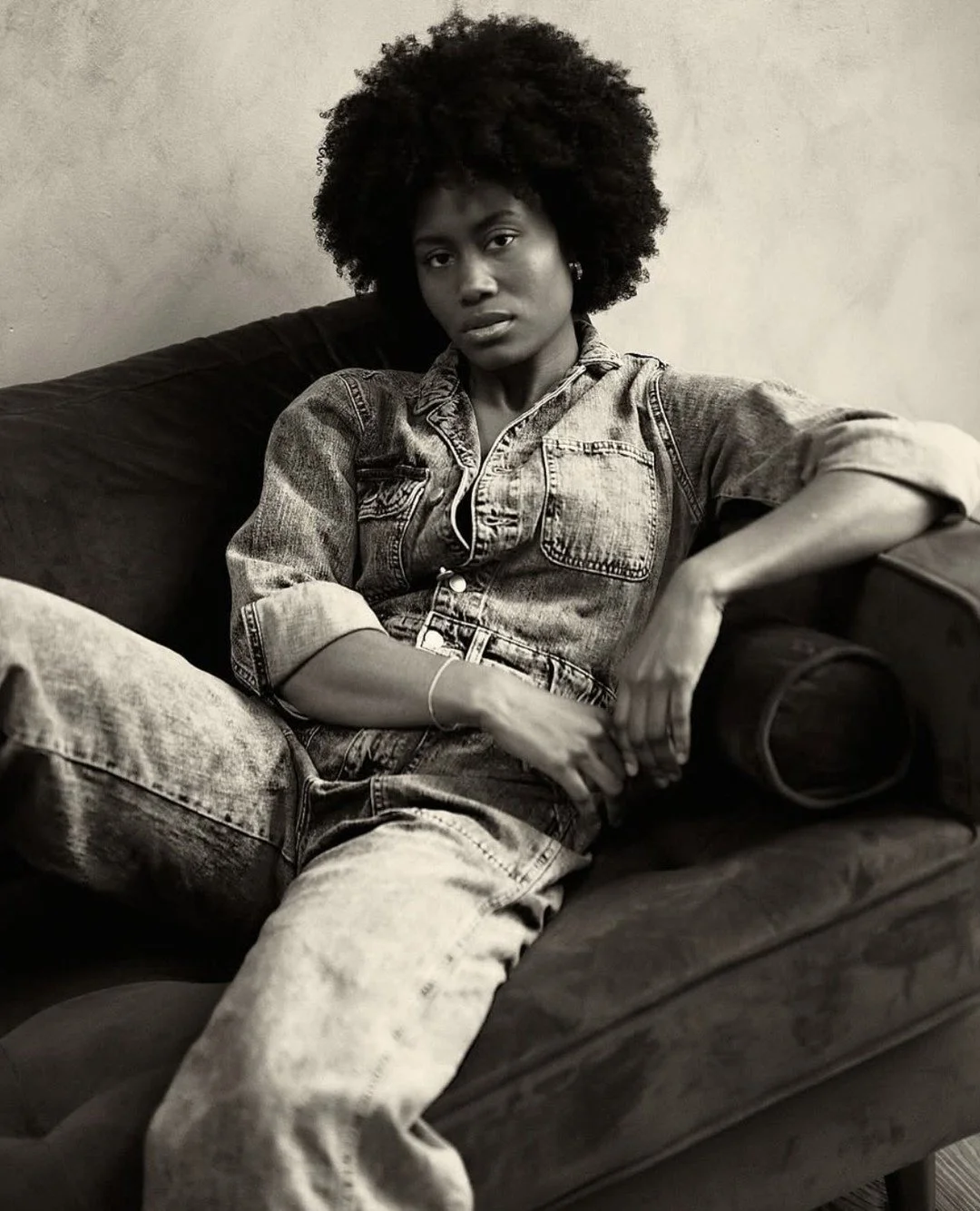 Black and white photo of a woman sitting on a sofa, wearing a denim jacket and jeans, with natural curly hair, looking directly at the camera.