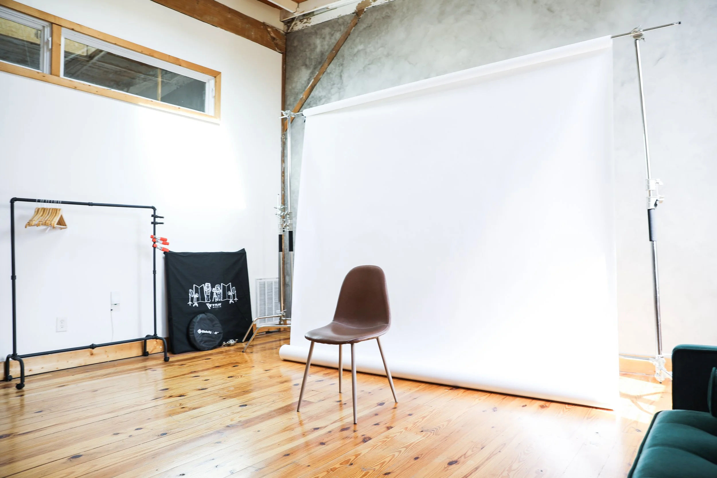 Photo of a photography studio with a white background and wooden floor. There is a brown chair in front of the backdrop, and a metal clothing rack on the left side of the image.