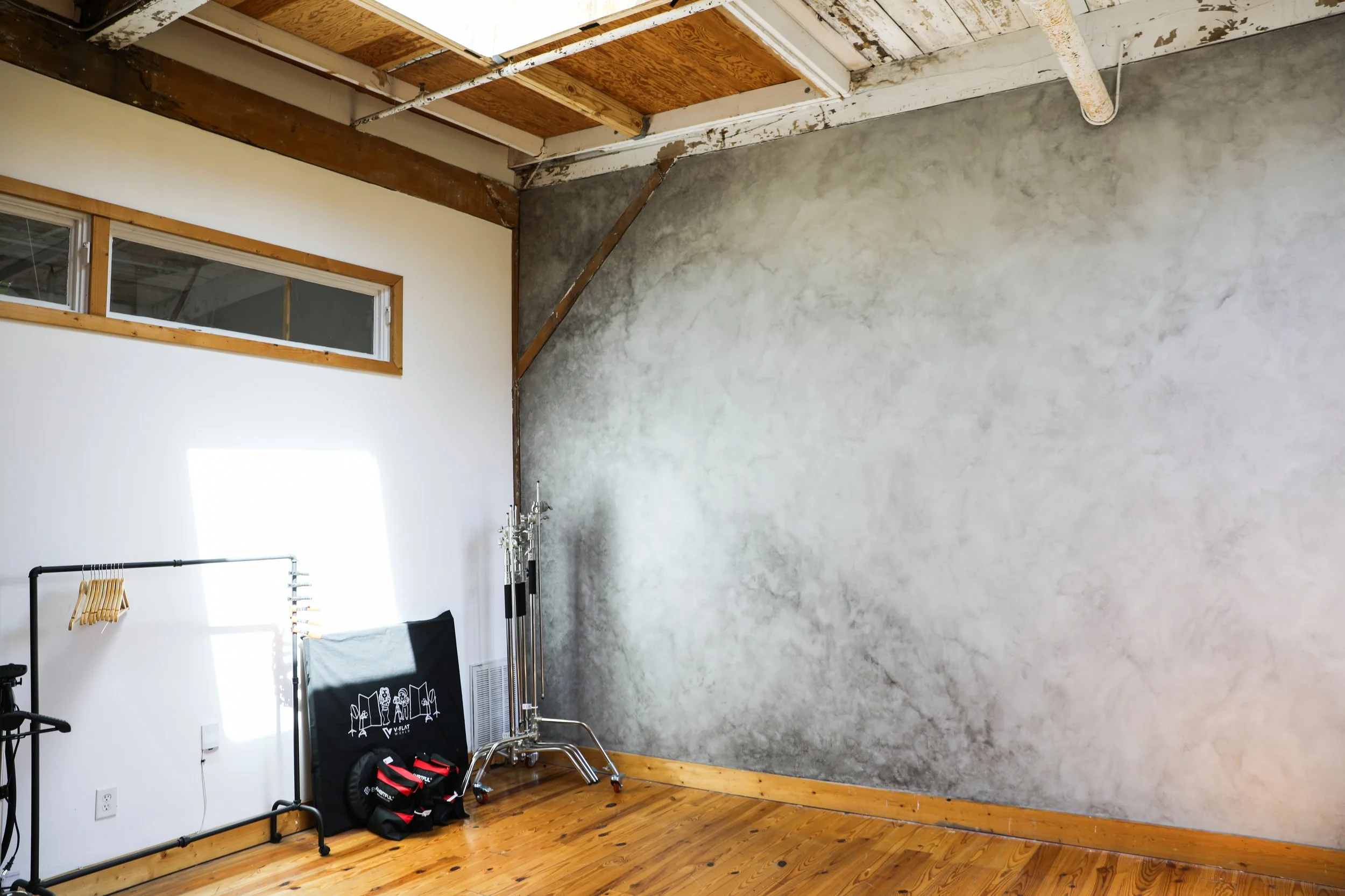 Interior of a room with a high ceiling, exposed wooden beams, a window high on the wall, a white wall with minimal decor, and a large unfinished gray wall, hardwood flooring, and some lighting and equipment on the floor.