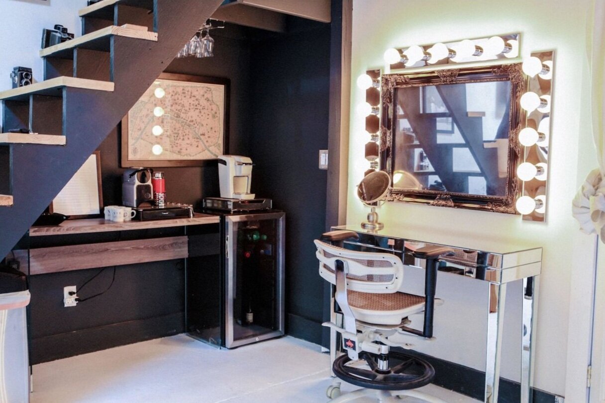 A modern room with a vanity mirror surrounded by lights, a makeup chair, a wall with a staircase, and a small bar area with coffee-making appliances and a mini-fridge.