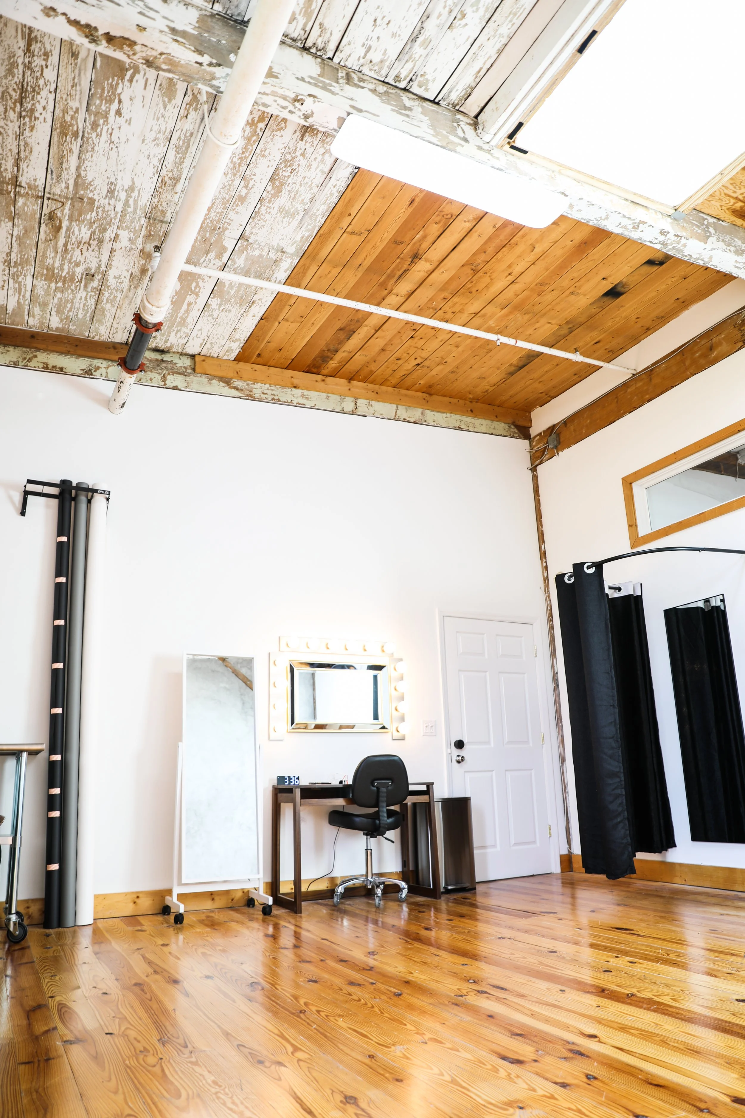 Interior of a room with a wooden floor, white walls, a white door, a black curtain, a black swivel chair at a small wooden desk with a mirror and vanity lights, a free-standing mirror, and rolled-up backdrops or fabric. The ceiling has a mix of expos