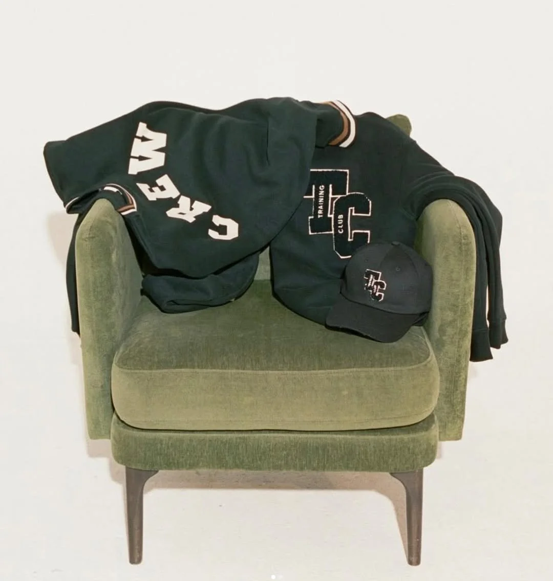 Green upholstered chair with a black sweatshirt, cap, and jacket draped over it, all featuring the 'GC' logo and branding.