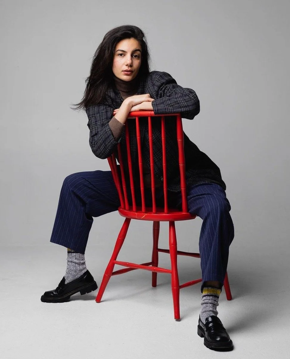 A young woman in a plaid blazer, dark turtleneck, and striped pants sits on a red chair. Her dark hair is loose, and she has a neutral expression. The background is plain gray.