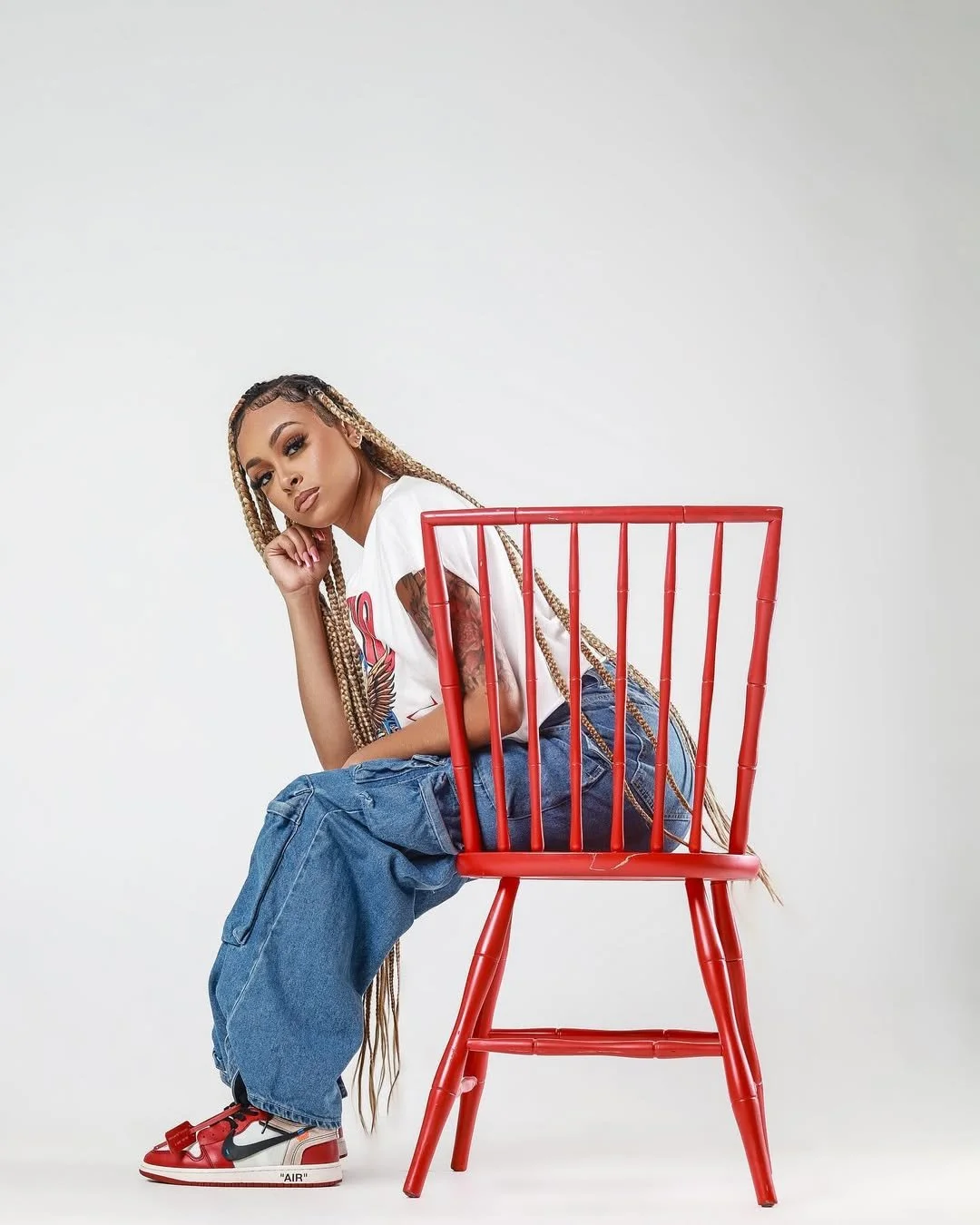 A woman with long, blond box braids sitting on a red chair with a white background. She has tattoos on her upper arm, is wearing a white T-shirt, baggy blue jeans, and red and white Nike sneakers.