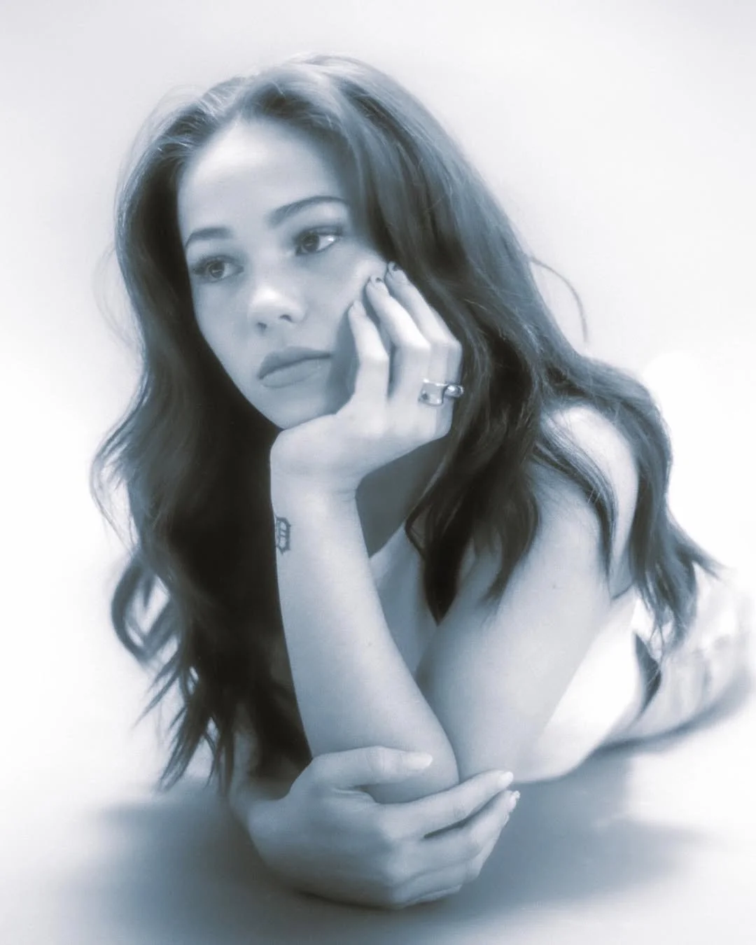 A black-and-white photo of a woman with long wavy hair resting her face on her hand, looking thoughtfully to the side.