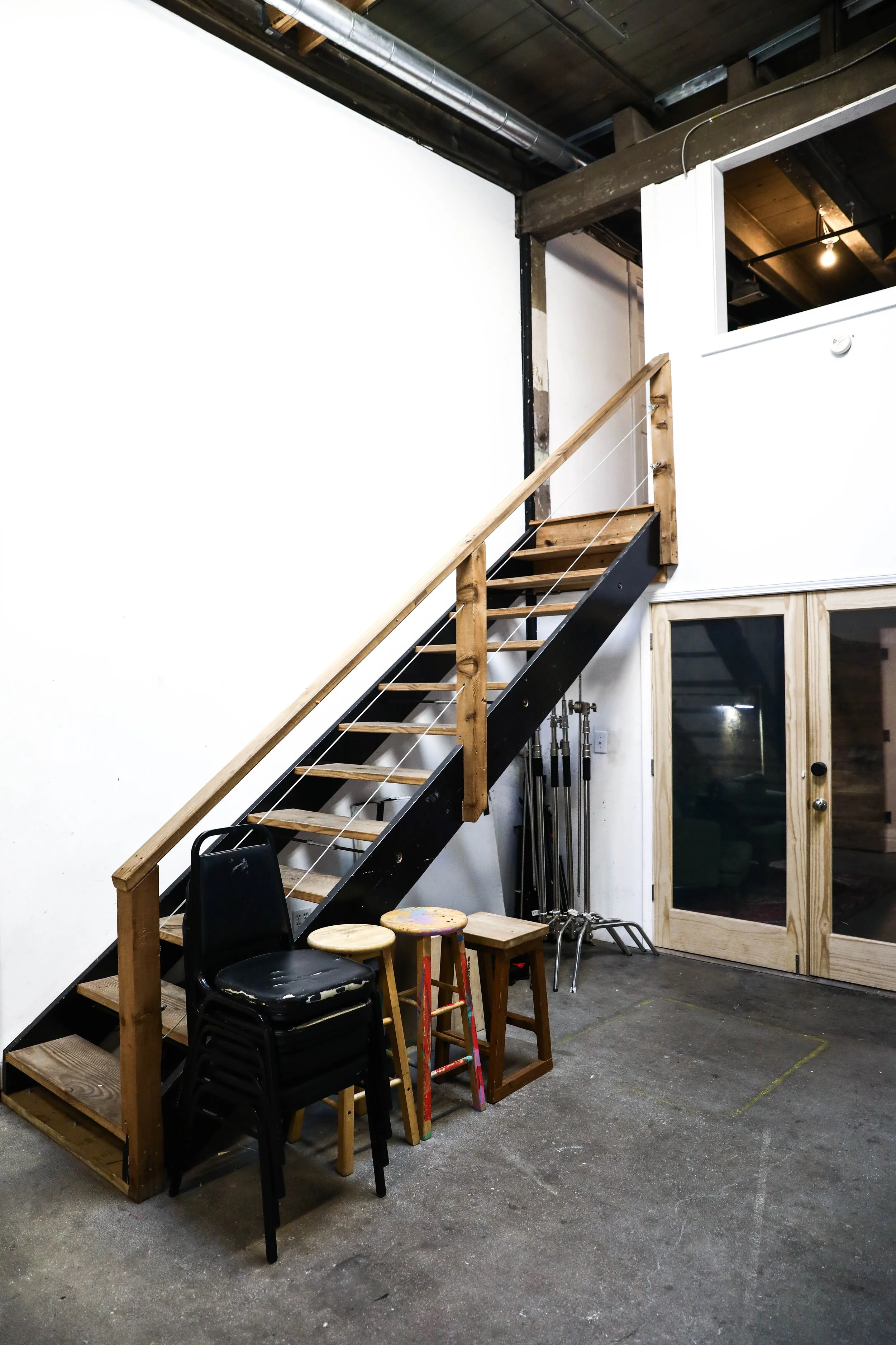 Interior of a room with a staircase leading to an upper level, stacked chairs, wooden stools, and gym equipment against a white wall.