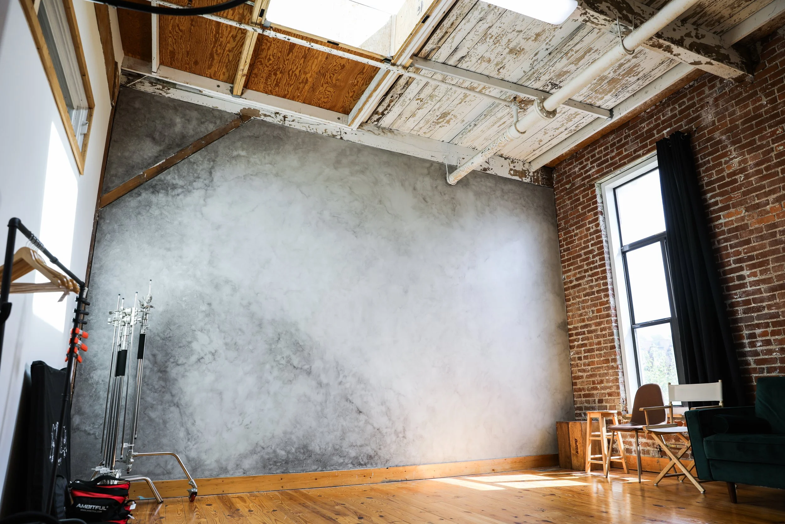 An empty room with a weathered wall painted gray, exposed brick wall, large window with black curtains, and a wooden floor. There are some chairs, a small wooden table, and equipment in the corner.