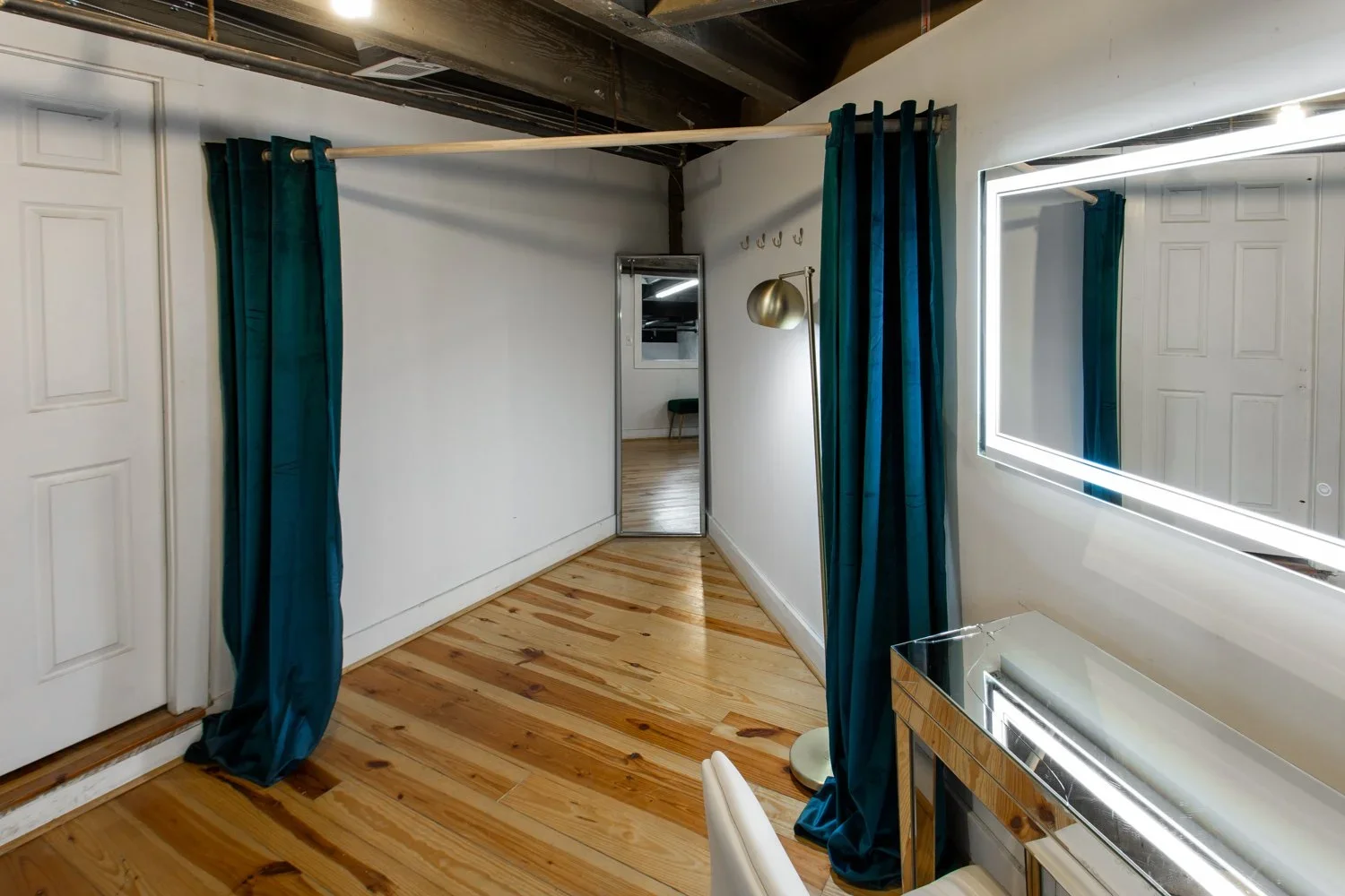 A fitting room with a mirror, blue curtains, a wooden floor, and a bathroom door in the background.