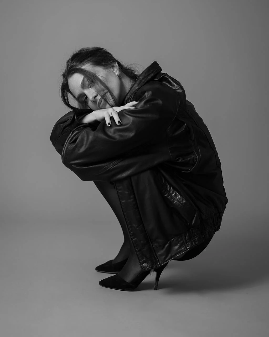 A woman with dark hair, wearing a black leather jacket, black high heels, and dark makeup, crouching with arms wrapped around her knees and eyes closed in a studio portrait shot.