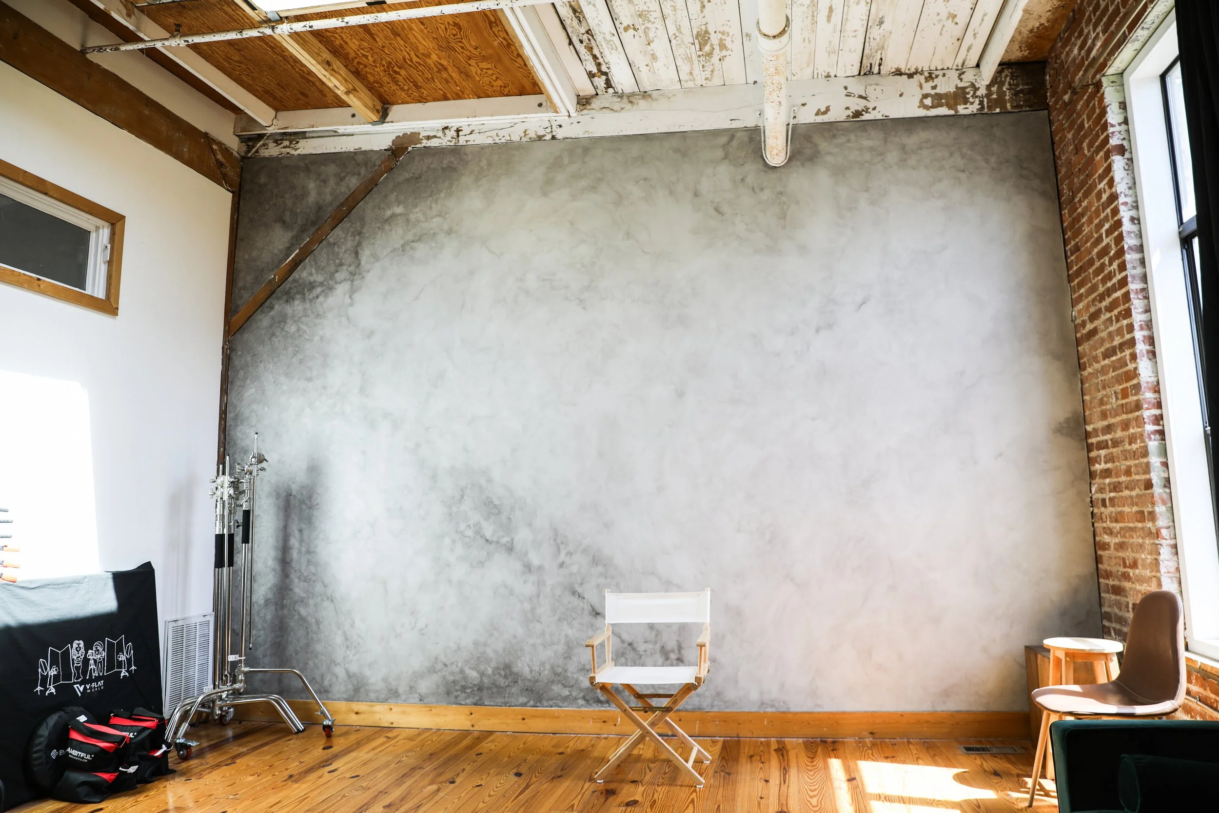 An empty room with a polished wood floor, a large gray wall, an exposed brick wall, and a high ceiling with unfinished wooden beams. There are two chairs and some equipment in the corner.