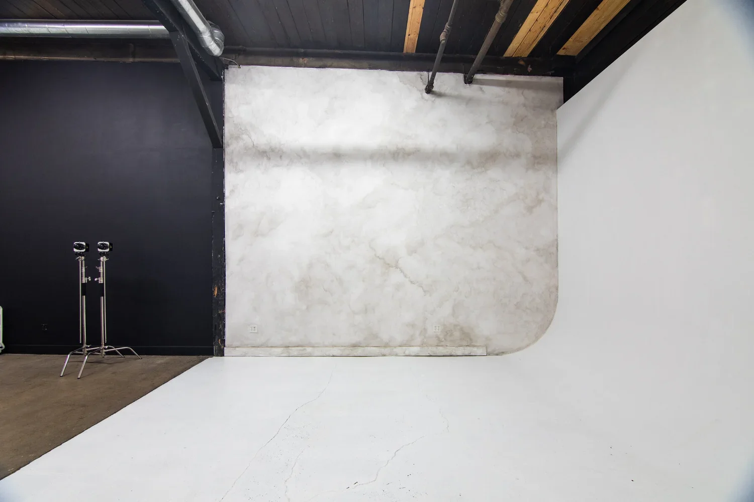 Empty room with white ceiling and wall, concrete wall with a rounded corner in the back, black wall with pipes on the left, two tripods next to each other on the left side, and electrical outlets on the concrete wall.