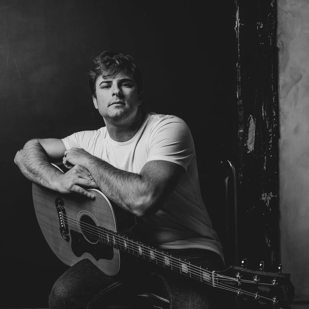 Black and white photo of a man in a white t-shirt sitting with his arms on an acoustic guitar against a dark wall.
