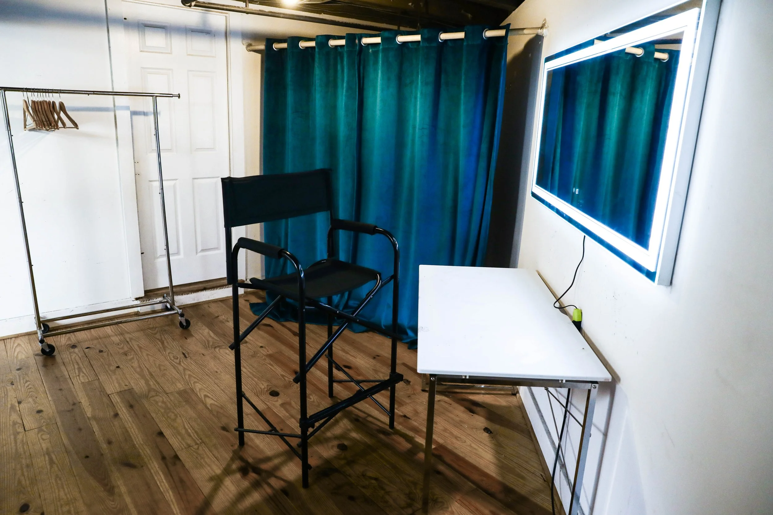 Empty room with a folding chair, a white table, a clothing rack with hangers, a mirror with LED light, and teal curtains.
