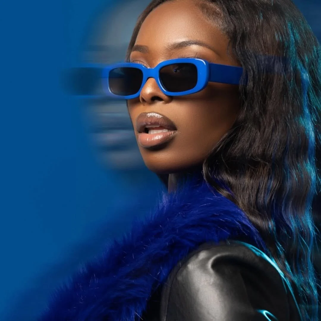 A woman with dark wavy hair wearing blue 3D glasses and a blue furry coat, looking to the side against a blue background with a motion blur effect.