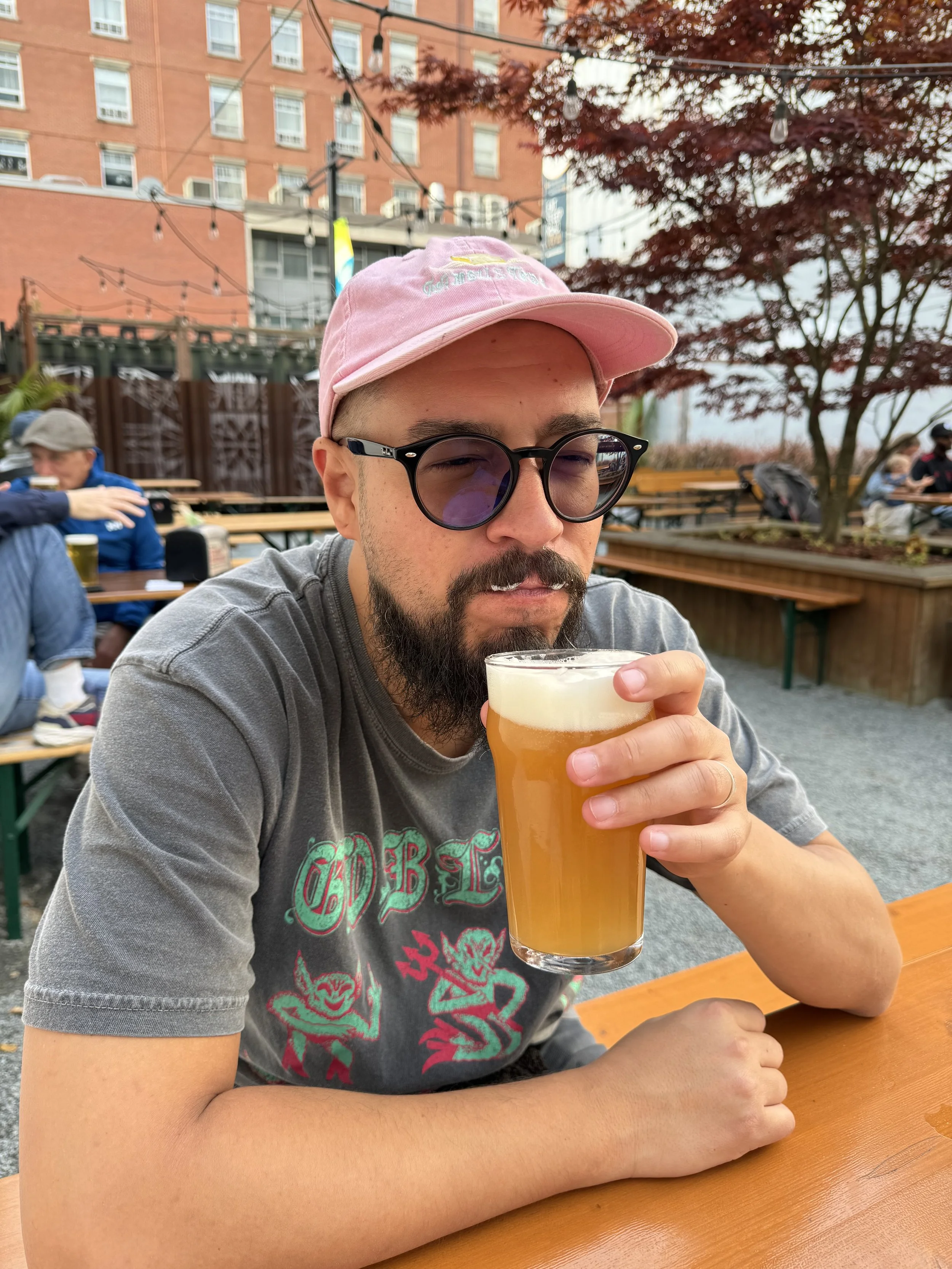 Neil is sitting at a beer garden drinking a beer with foam in his moustache