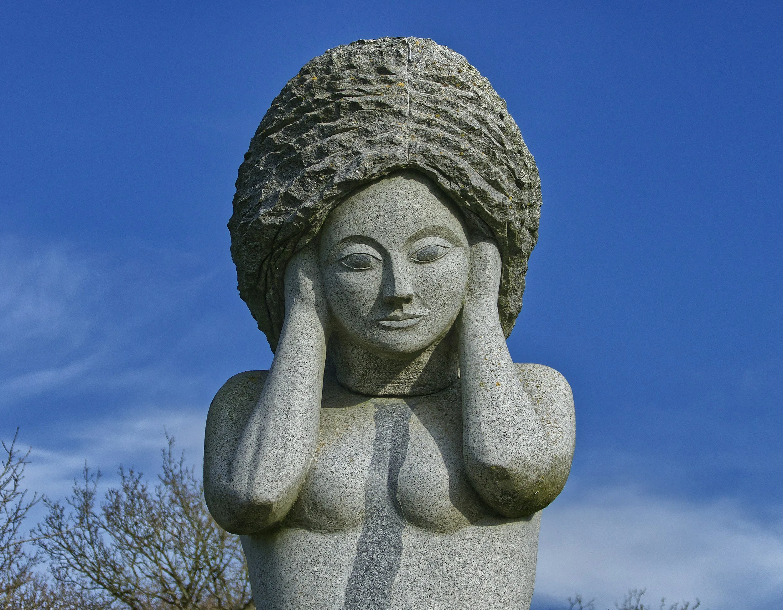 Stone sculpture of a woman with a large, textured hairstyle, holding her face with her hands, set against a blue sky with some tree branches below.