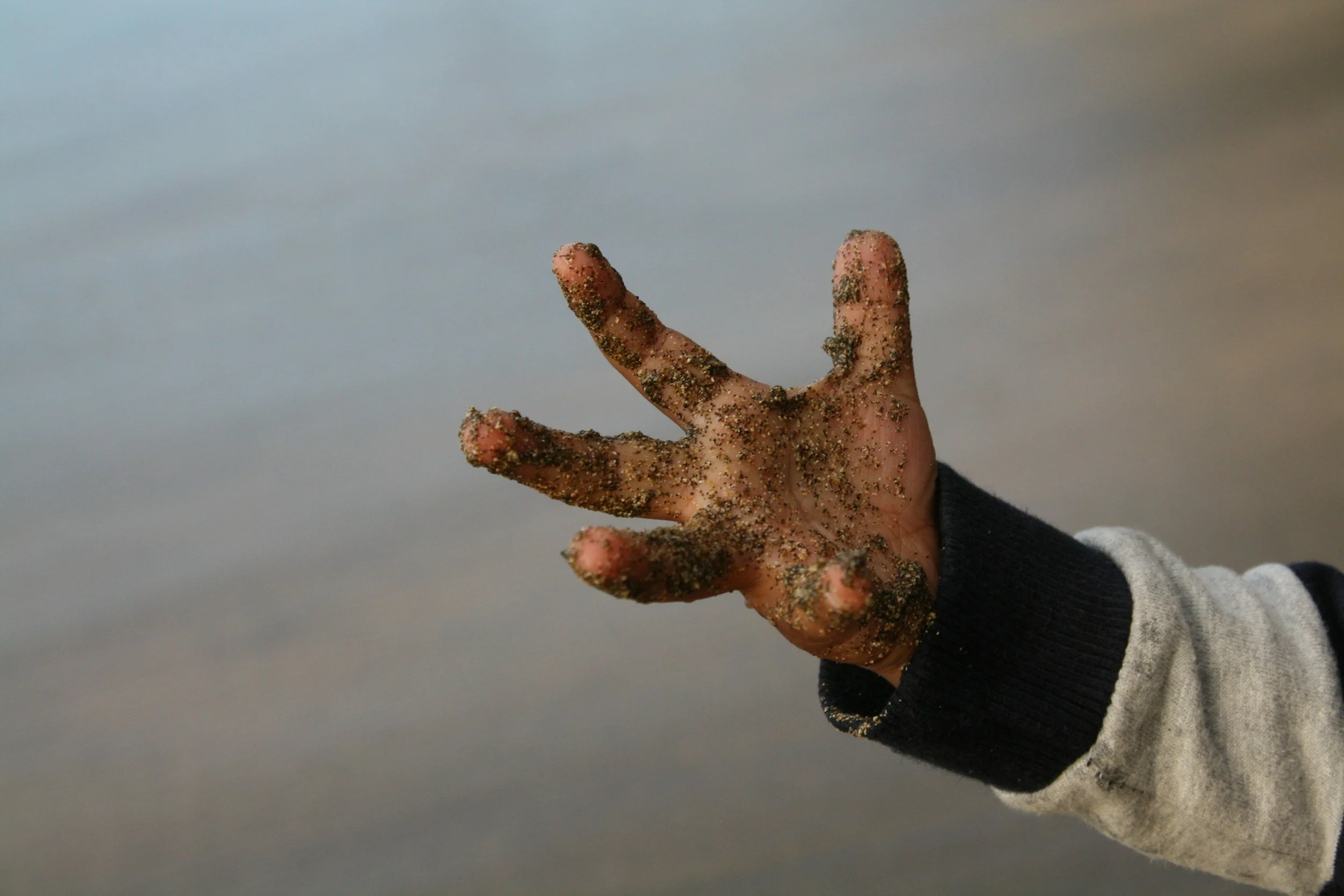 A human hand covered in sand, reaching out against a blurred background of water or sky.