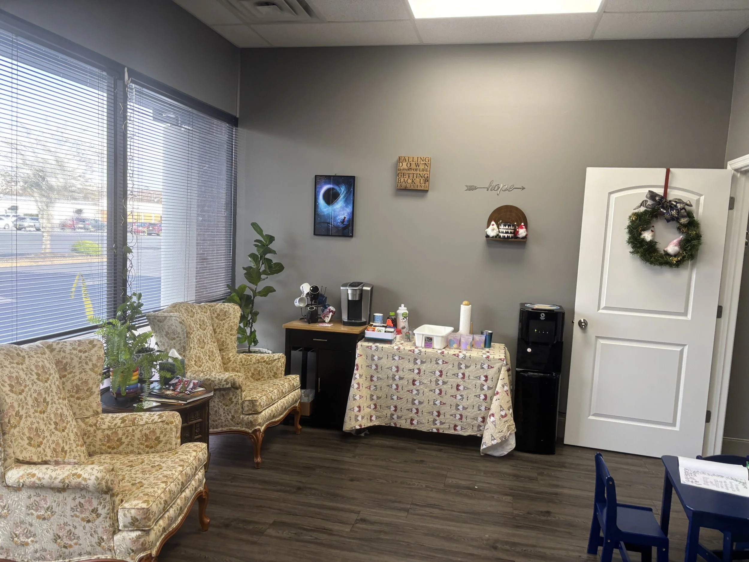Interior office with vintage style chairs, a table with dishes and supplies, a water cooler, and holiday decorations including a wreath on the door, Christmas figurines, and wall signs.