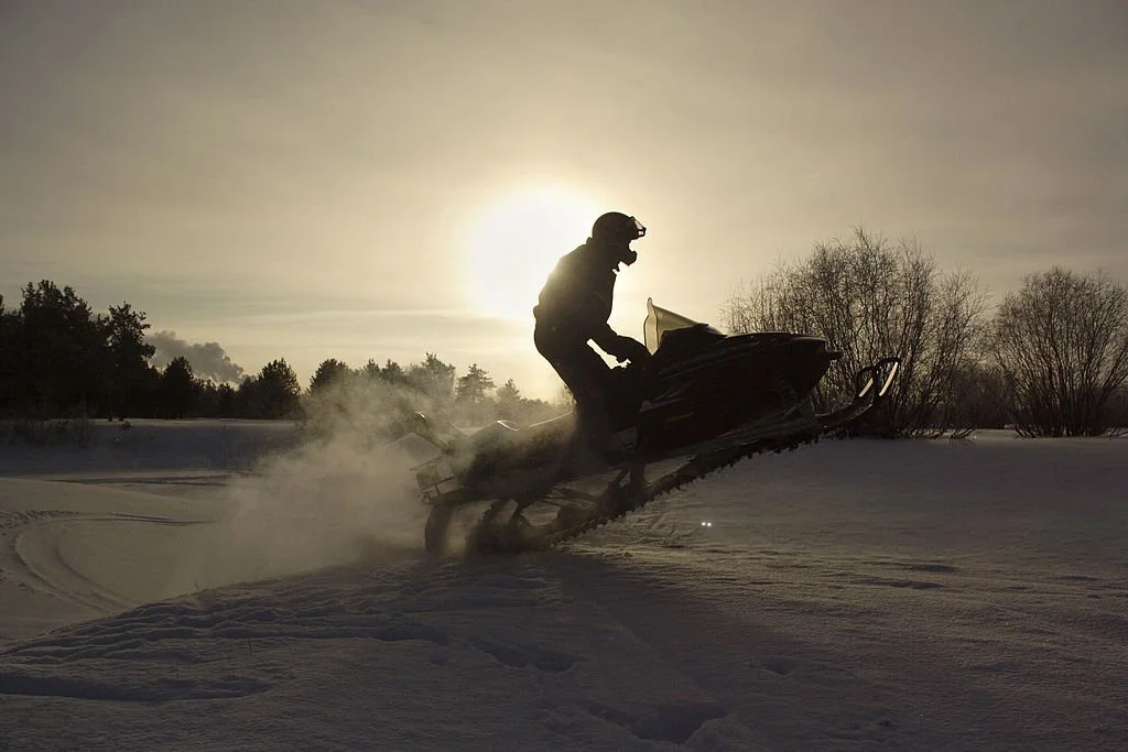 A person riding a snowmobile through snow with the sun setting or rising in the background, creating a silhouette and kicking up snow after filling up with VentPro