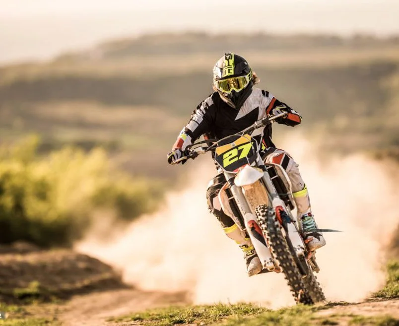 A motocross rider in black and white gear with a yellow helmet and goggles, riding a dirt bike numbered 27 on a dusty trail in a natural outdoor setting.