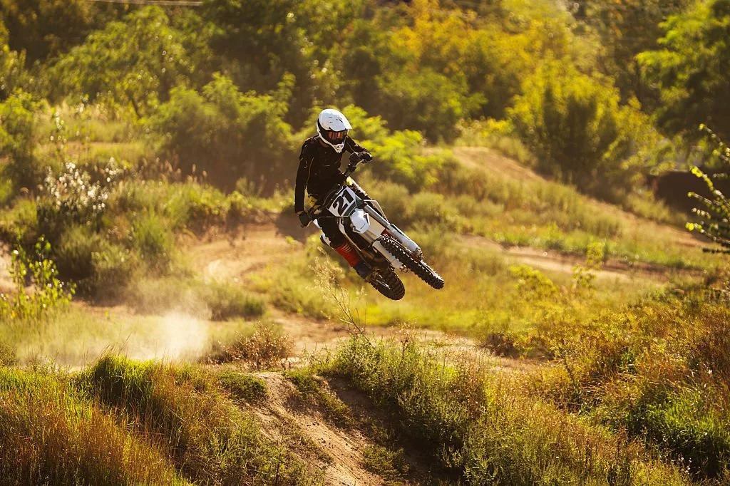 A person riding a dirt bike and jumping over a trail in a grassy, wooded area during daytime.
