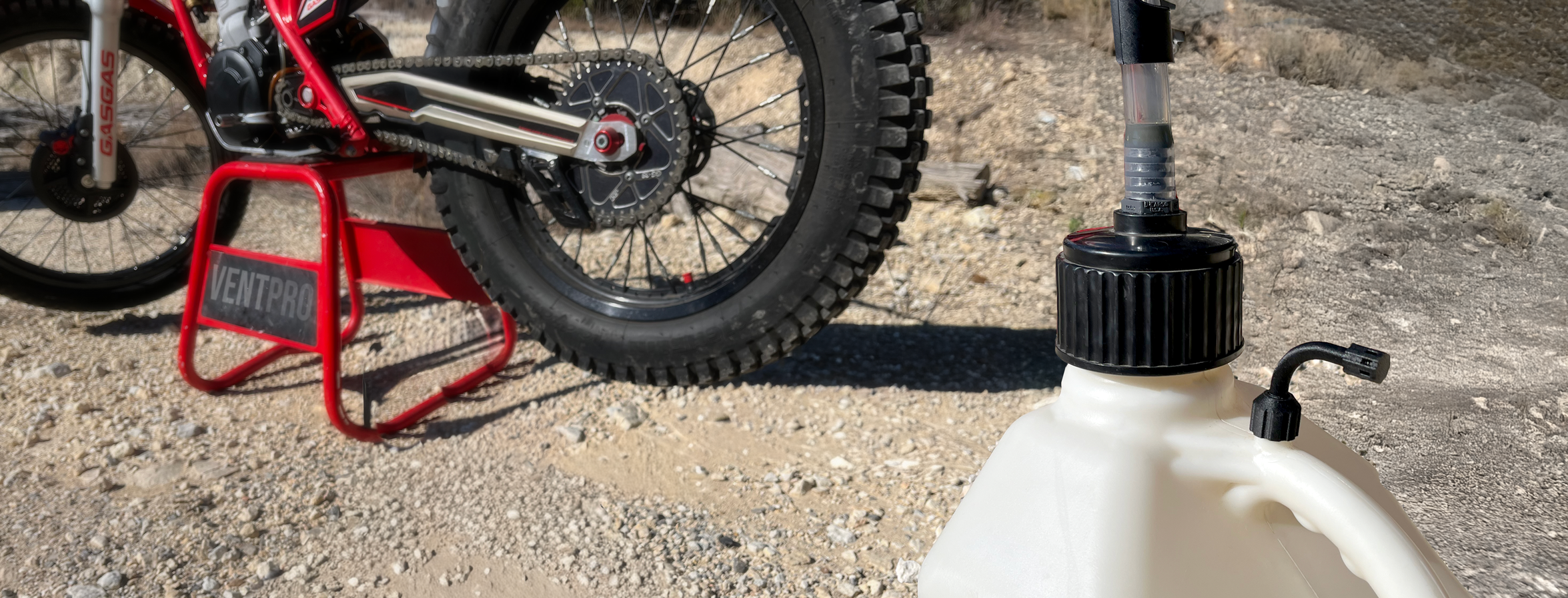 Close-up of a dirt bike on a red stand with a large knobby rear tire. In the foreground, there is a white plastic container with a black screw-on cap and a tube attached.