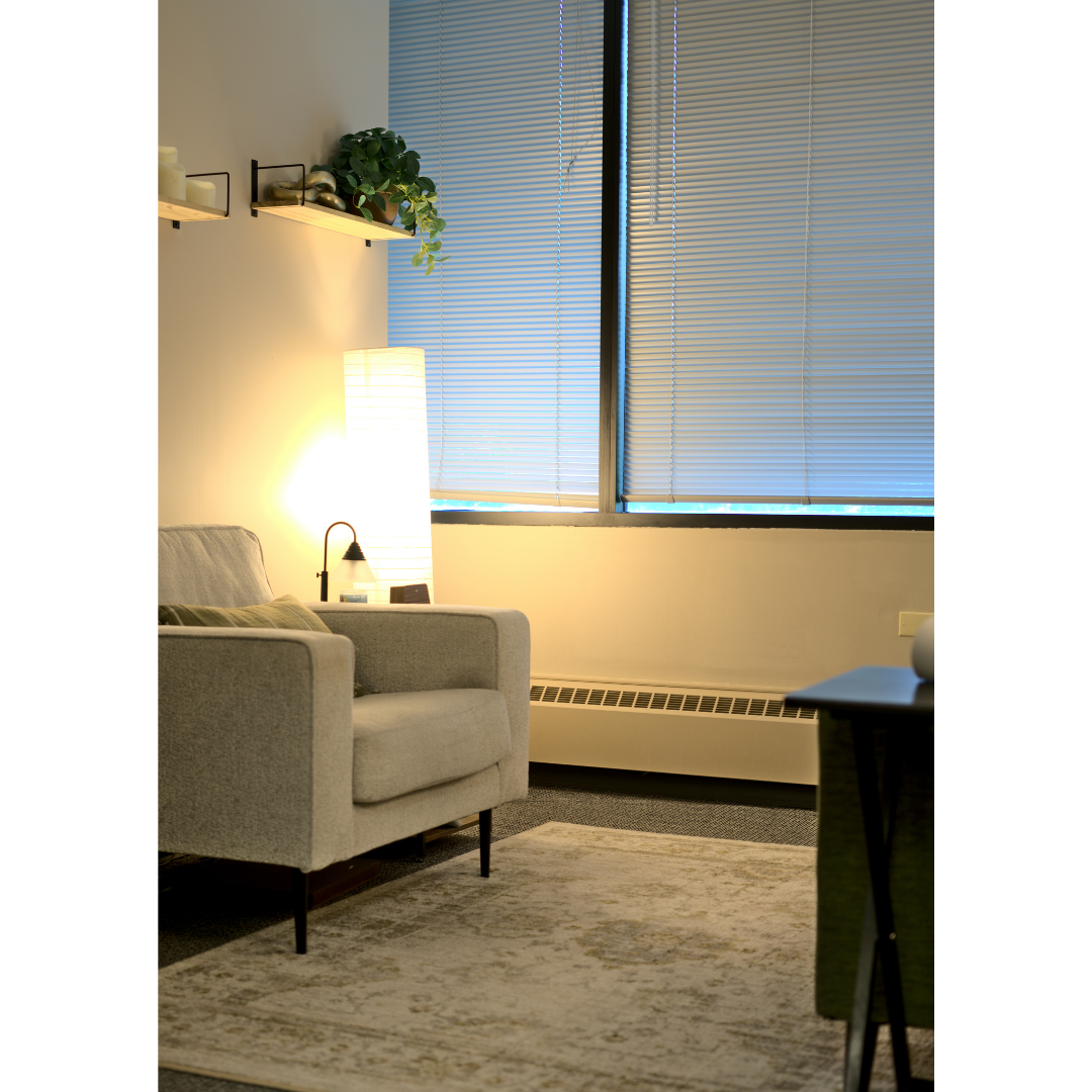 A cozy living room corner with a beige armchair, a tall white floor lamp, a small table with a lamp and other items, two shelves with decorative objects and a potted plant, and window blinds covering large windows.