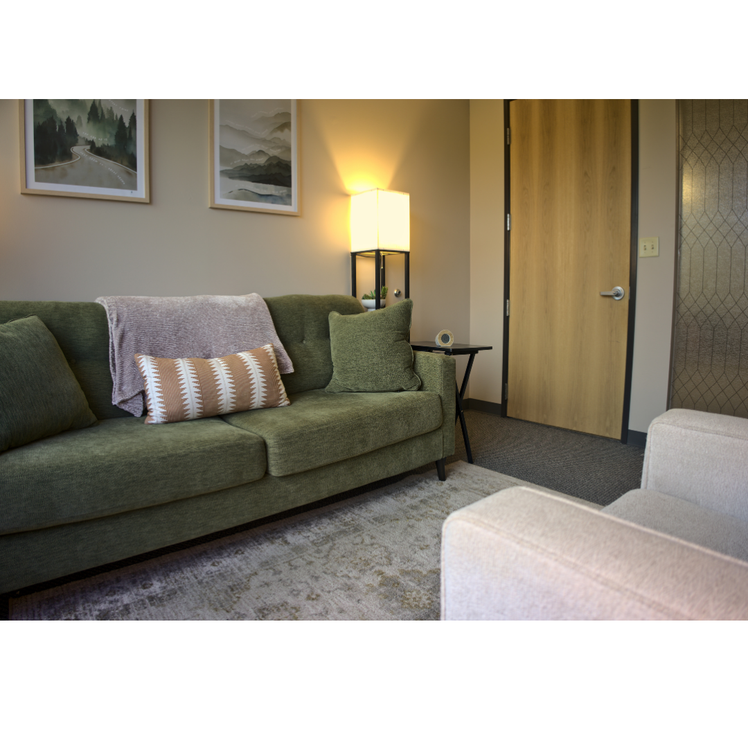 Living room with a green sofa, beige arm chair, colorful artwork on the wall, standing lamp, side table, and a closed wooden door.