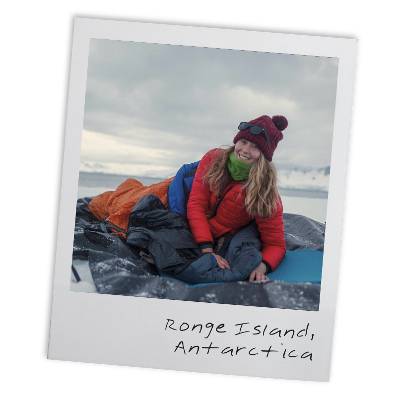 A woman in outdoor gear, including a red jacket, green neck warmer, maroon beanie with pom-pom, and sunglasses, smiling while lying on a sleeping bag on a cloudy, icy landscape in Ronge Island, Antarctica.