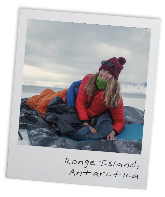 A woman lying on a black inflatable boat on the beach in Antarctica, wearing a red jacket, purple beanie, green scarf, and sunglasses on her beanie, smiling at the camera with snow and ice in the background.