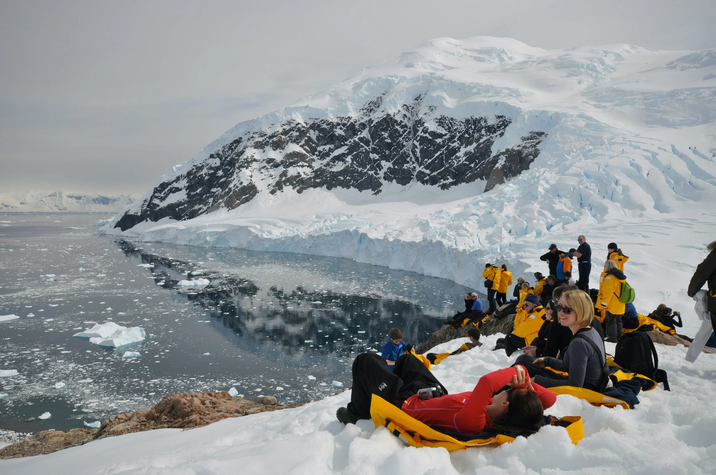 Tourists in yellow jackets resting on snow-covered terrain near icy waters and a large glacier in Antarctica.