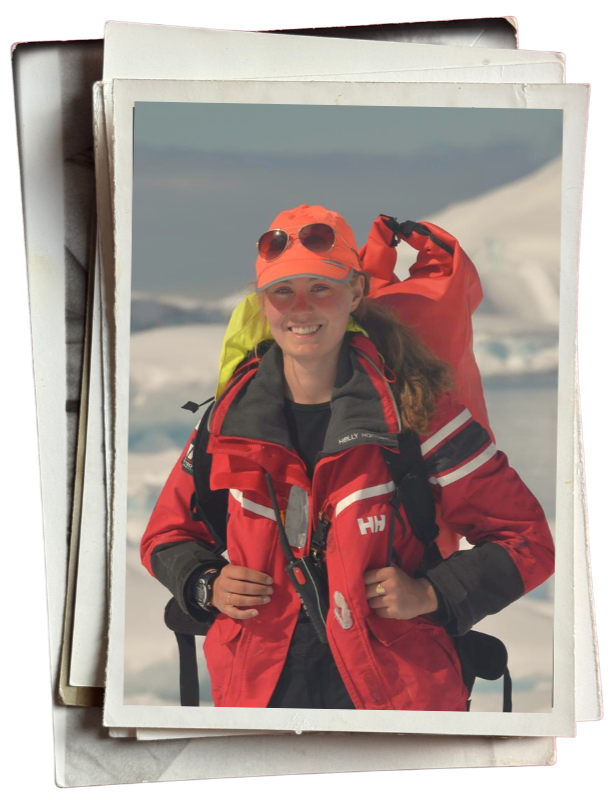 A young woman with long hair, wearing sunglasses, an orange cap, and a red jacket, smiling while hiking in a snowy, icy landscape.