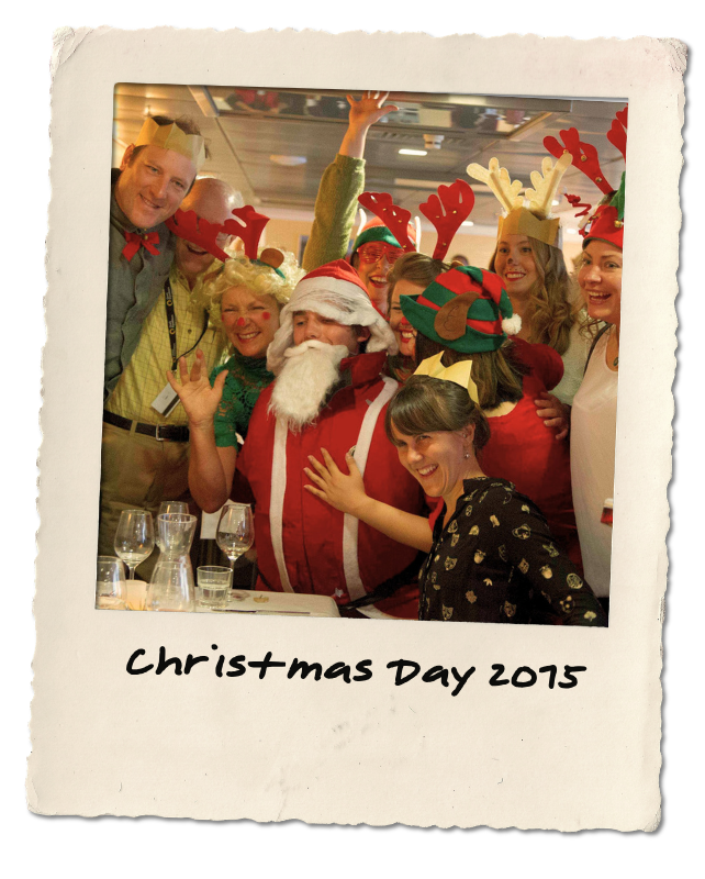 Group of people celebrating Christmas, dressed in festive costumes including reindeer antlers, Santa hat, and elf hat, gathered around a person dressed as Santa Claus. The photo is labeled 'Christmas Day 2015' at the bottom.