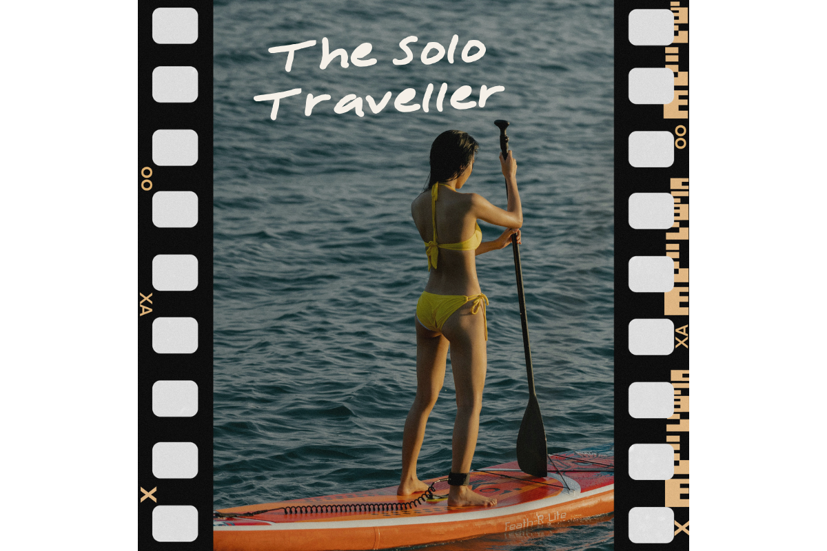 A woman in a yellow bikini stands on a paddleboard holding a paddle, facing away from the camera on a body of water, with carpentry markings and the text 'The Solo Traveller' overlayed.