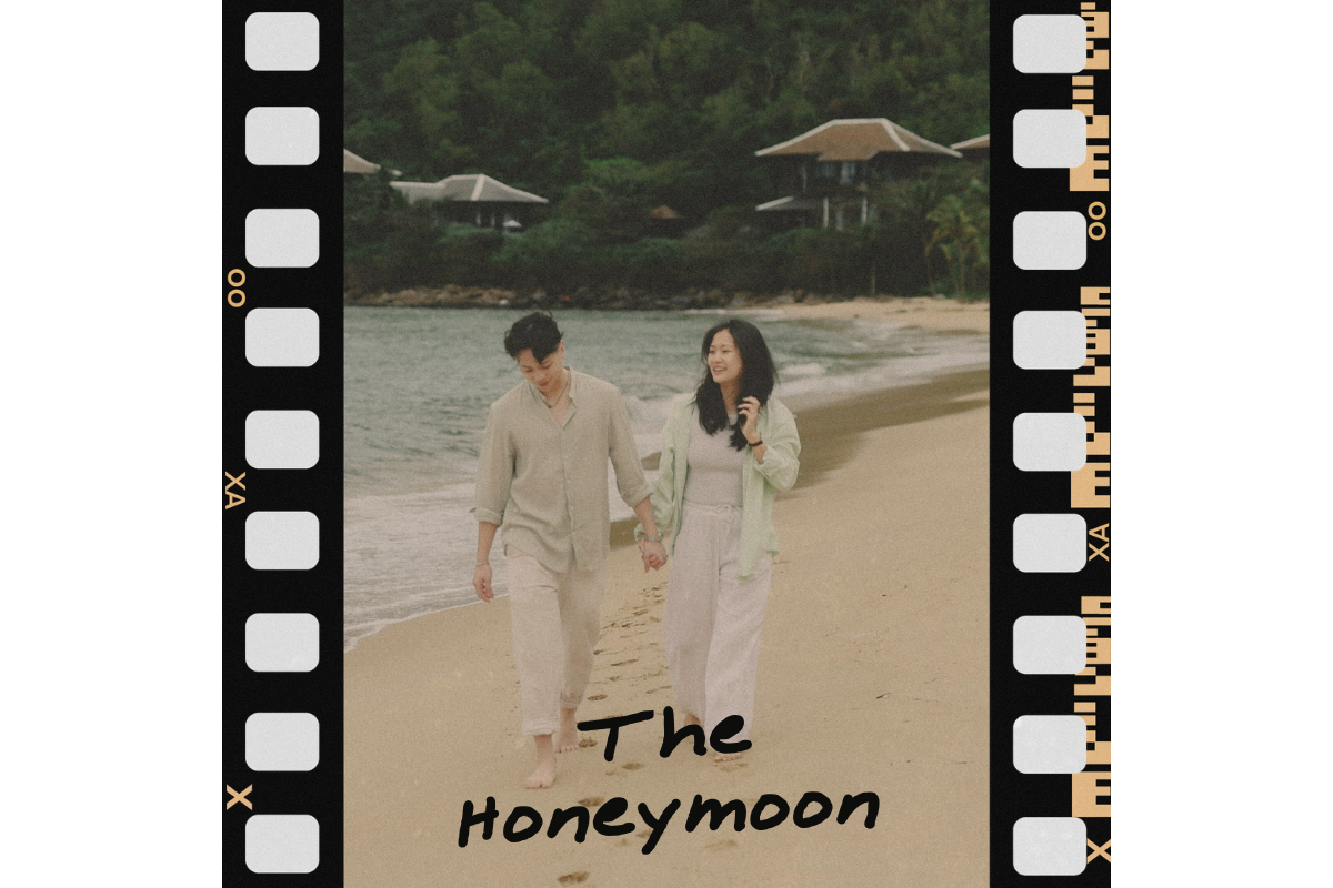 A couple walking hand in hand along a beach, holding hands, with the ocean and greenery in the background, and the text 'The Honeymoon' at the bottom.